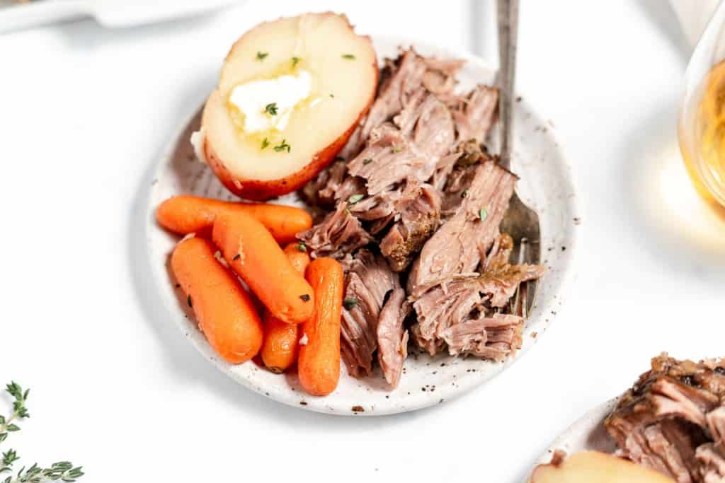 A plate of sliced roast beef with baby carrots and a halved baked potato topped with butter, garnished with thyme, on a white surface.