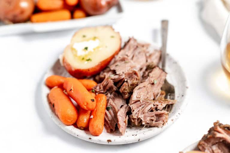 A plate of pot roast with carrots and a half potato topped with butter, accompanied by a glass of white wine.