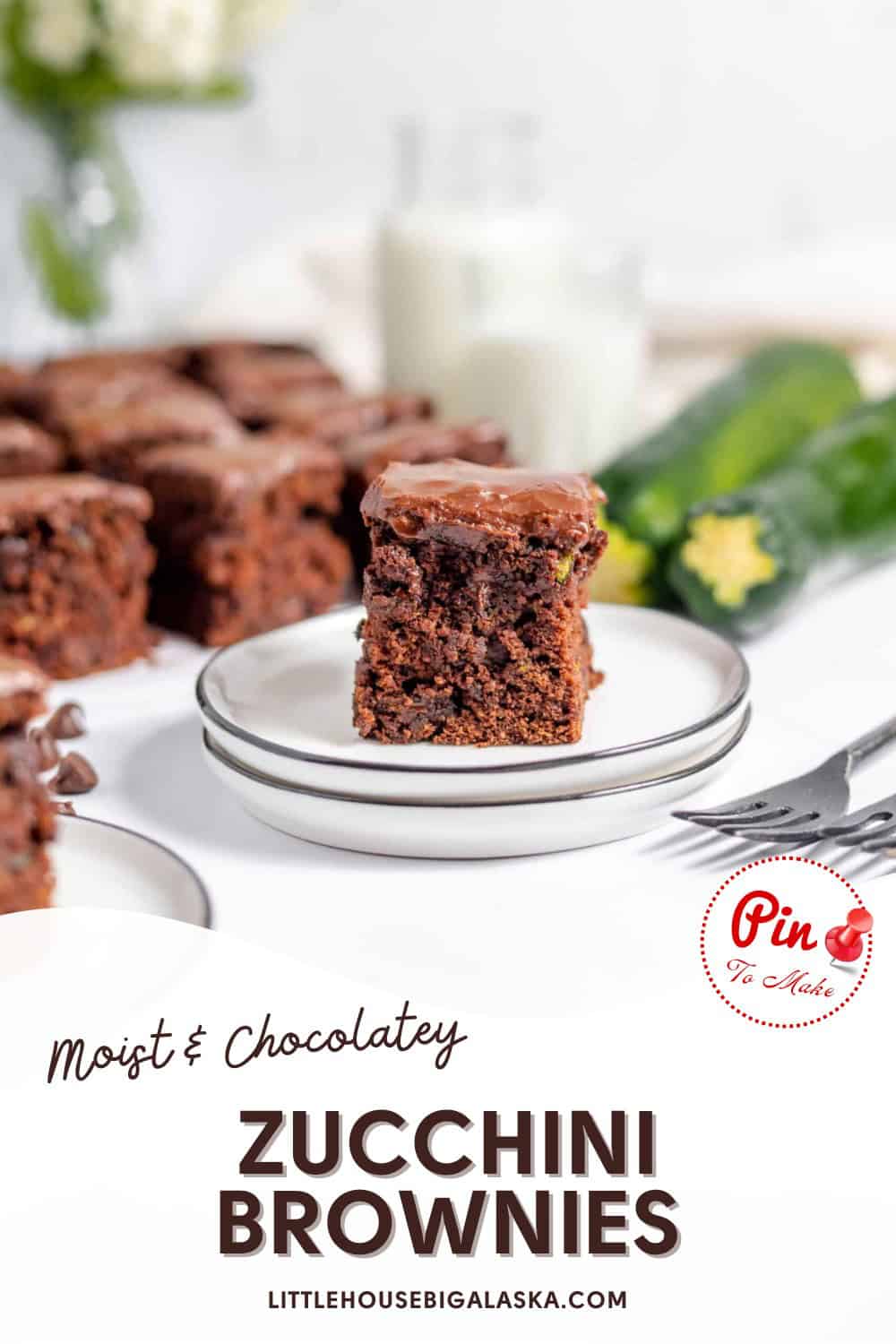 A stack of chocolate zucchini brownies on a white plate, with a larger plate of brownies, zucchini, and a glass of milk in the background. Text reads "Moist & Chocolatey Zucchini Brownies.