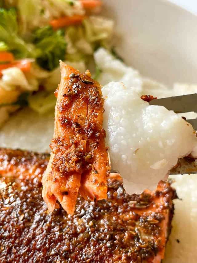 Close-up of a fork holding a bite of seasoned salmon and rice, with a portion of salad visible in the background.