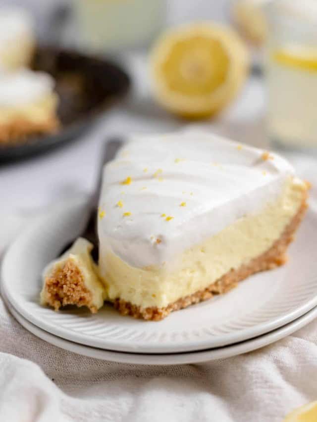 A slice of lemon cream pie sits on a white plate with a fork. The pie has a graham cracker crust, creamy lemon filling, and a layer of whipped topping garnished with lemon zest.