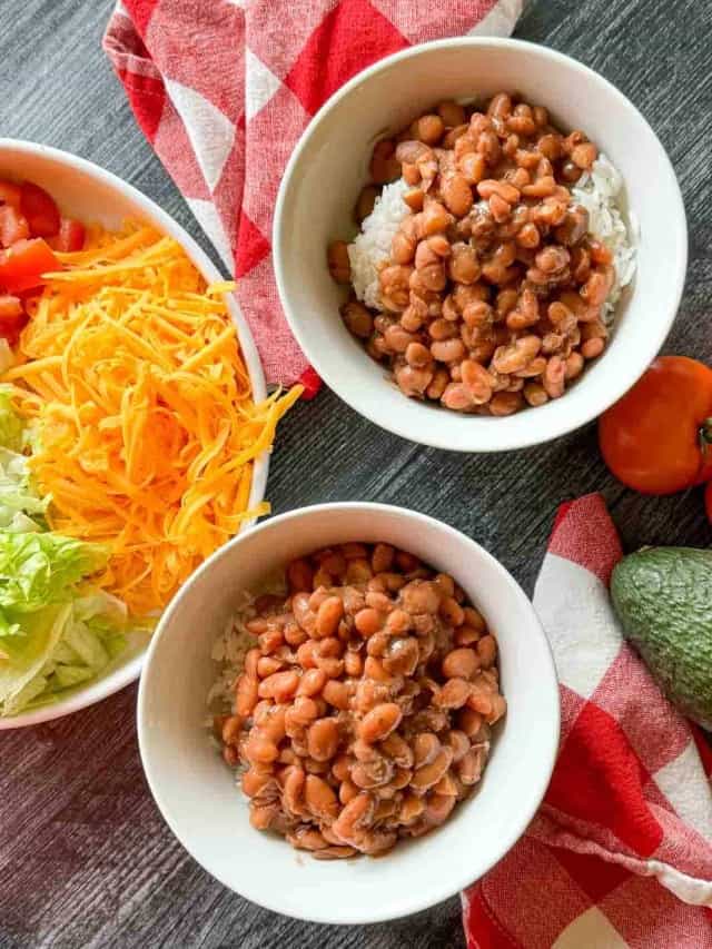 Two bowls of rice topped with beans are on a table next to a bowl of shredded cheese and chopped vegetables, including lettuce and tomatoes. An avocado and a tomato are placed nearby.