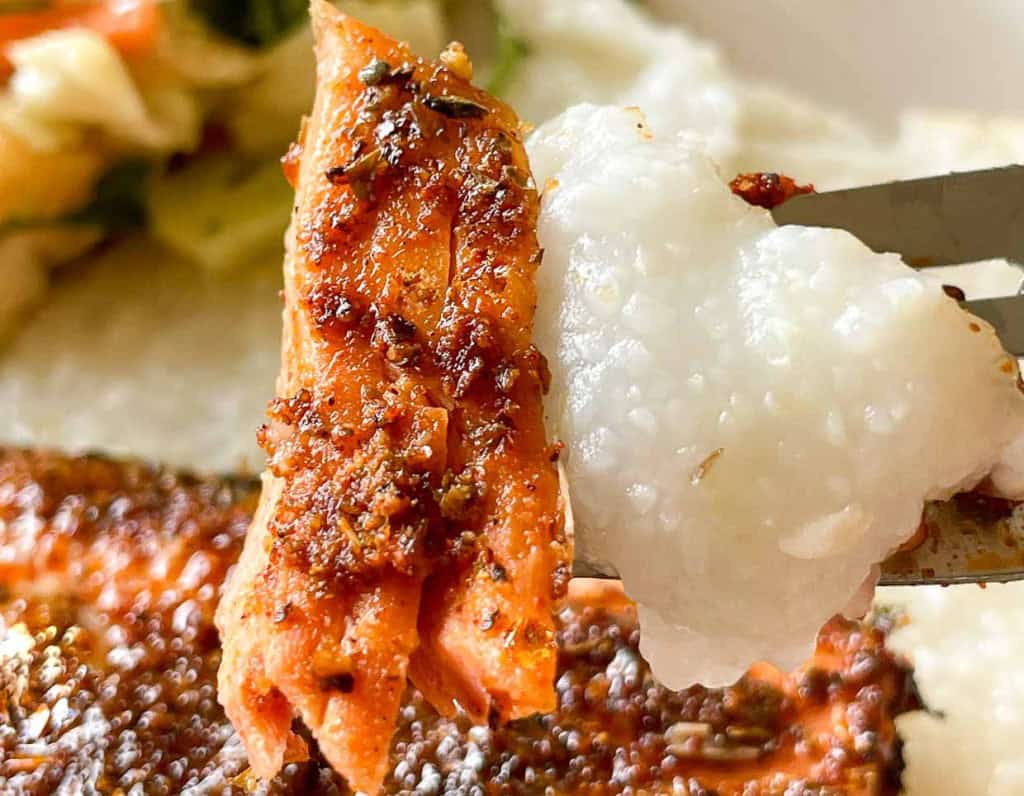 A fork holding a piece of seasoned, cooked salmon and a portion of white, creamy grits, with vegetables blurred in the background.