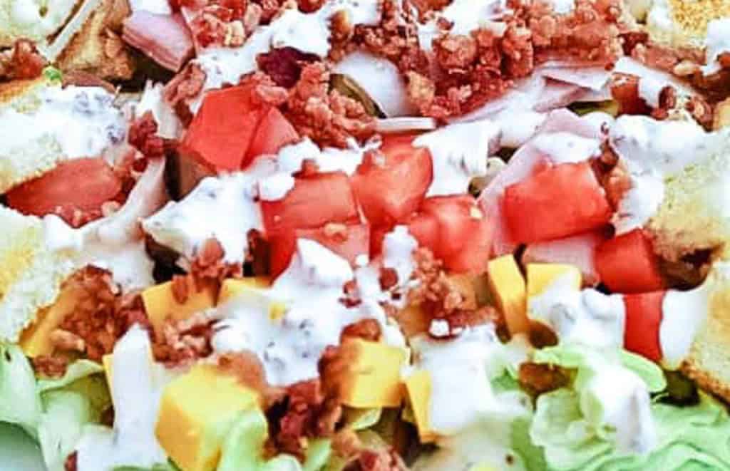 A close-up of a salad featuring diced tomatoes, shredded cheese, bacon bits, and croutons, all topped with a creamy dressing. Big salads we love to make for dinner like this one are perfect for any meal.