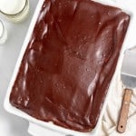 A rectangular chocolate sheet cake in a white dish, placed on a marble surface. A large knife with a wooden handle and a white cloth are on the right, with two glasses of milk on the left.