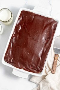 A rectangular chocolate sheet cake in a white dish, placed on a marble surface. A large knife with a wooden handle and a white cloth are on the right, with two glasses of milk on the left.