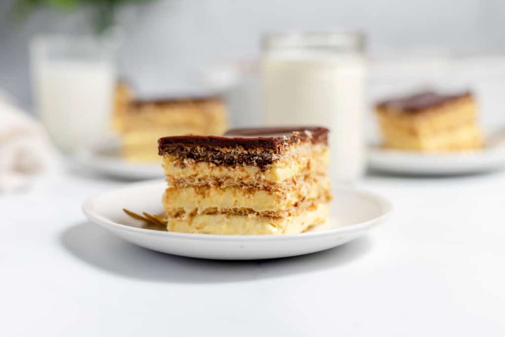 A slice of layered dessert with cream and chocolate on a white plate, with additional slices and glasses of milk in the background.