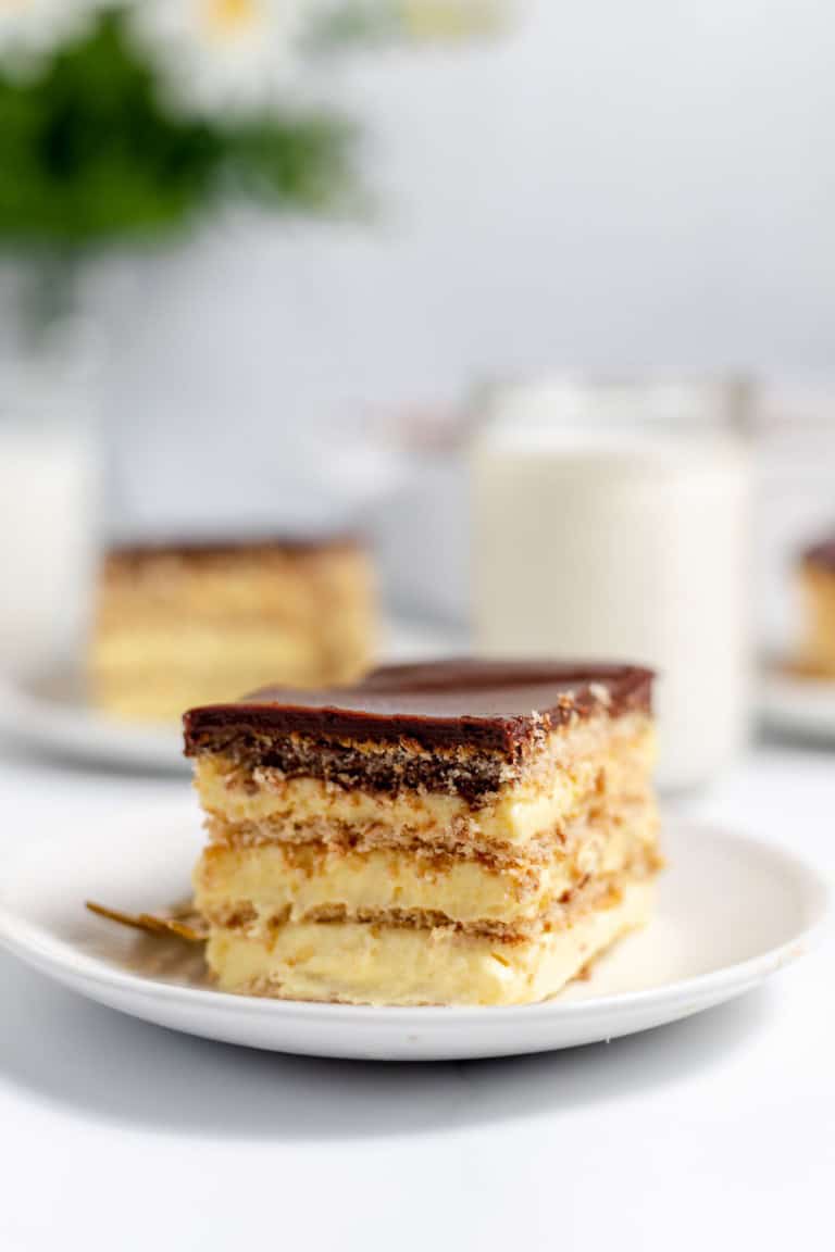 A slice of layered dessert with cream and chocolate topping on a white plate, with a glass of milk and another slice in the background.