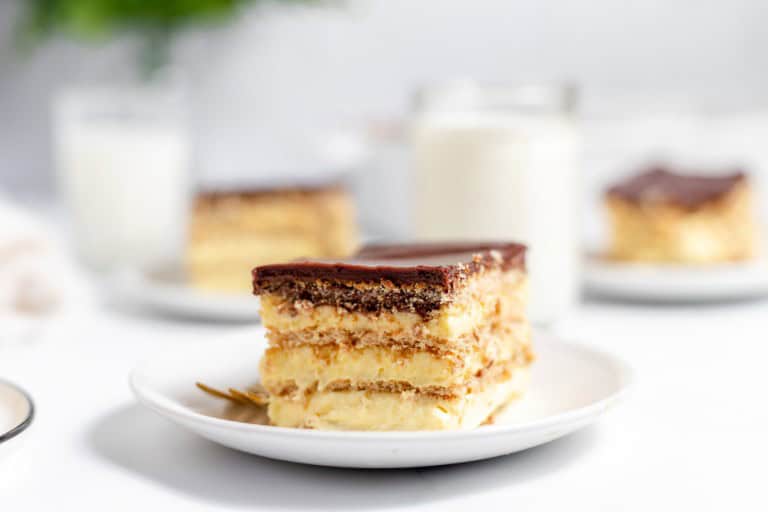 A slice of layered cake with chocolate topping on a white plate. Blurred background includes another piece of cake, a glass of milk, and a green plant.