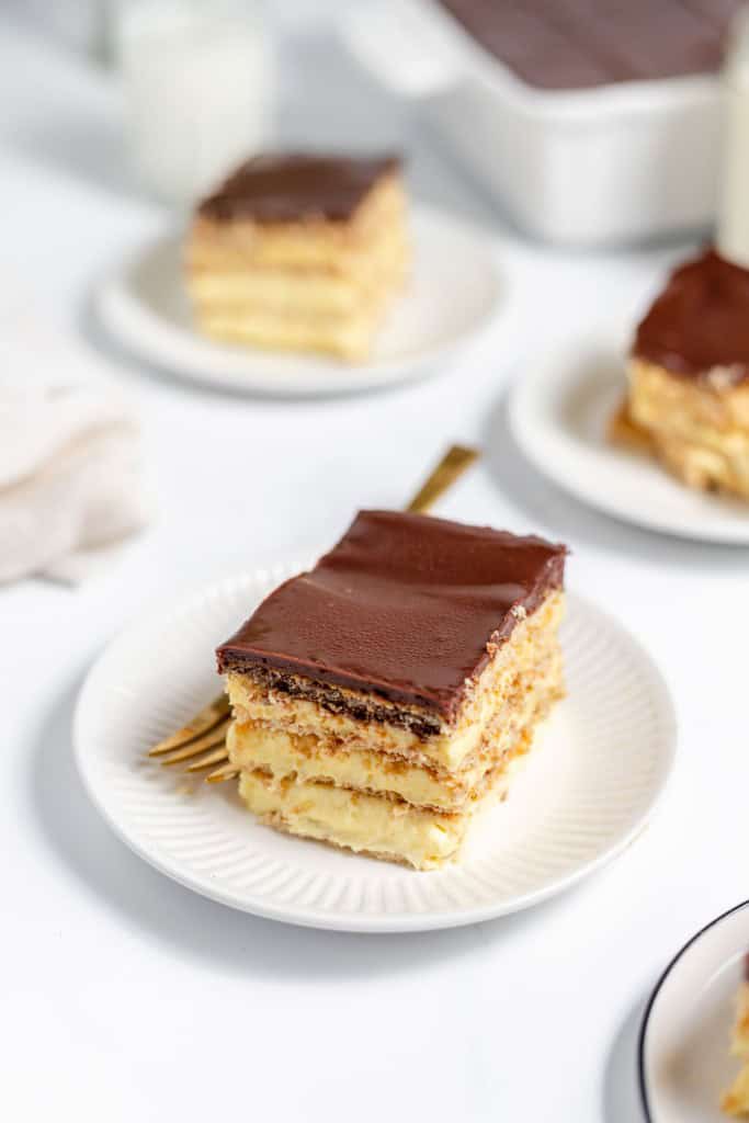 A slice of chocolate eclair cake on a white plate with a gold fork, with more slices in the background. The cake has layers of cream and a chocolate topping.