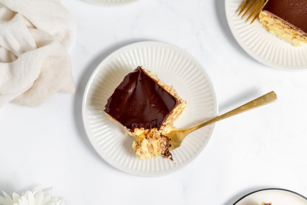 A slice of layered dessert with a chocolate glaze on a white plate, accompanied by a gold fork. The dessert appears partially eaten.