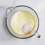 A glass pitcher containing a mixture of milk and a mound of dry ingredients, viewed from above, on a marble surface.