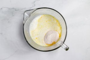 A glass pitcher containing a mixture of milk and a mound of dry ingredients, viewed from above, on a marble surface.