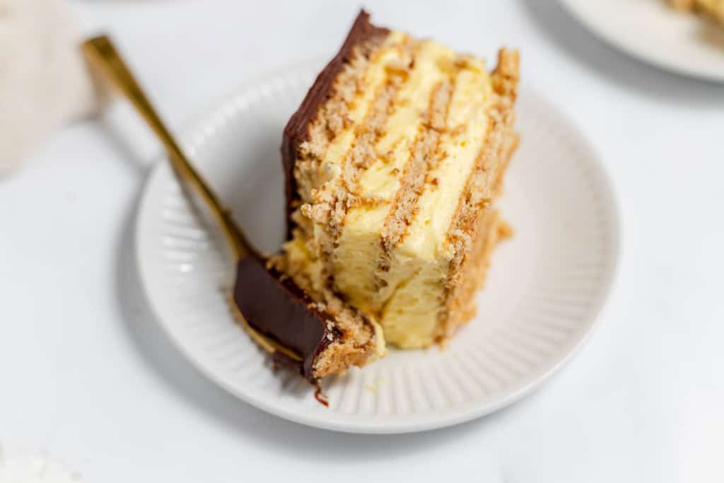 A slice of layered cake with chocolate topping and creamy filling on a white plate, accompanied by a fork with a piece of cake on its prongs.