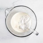 A glass measuring cup filled with cream and a heap of flour, placed on a white marble surface.