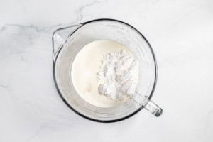 A glass measuring cup filled with cream and a heap of flour, placed on a white marble surface.