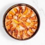 A round cake topped with sliced peaches and dusted with sugar in a metal baking pan on a marble countertop.