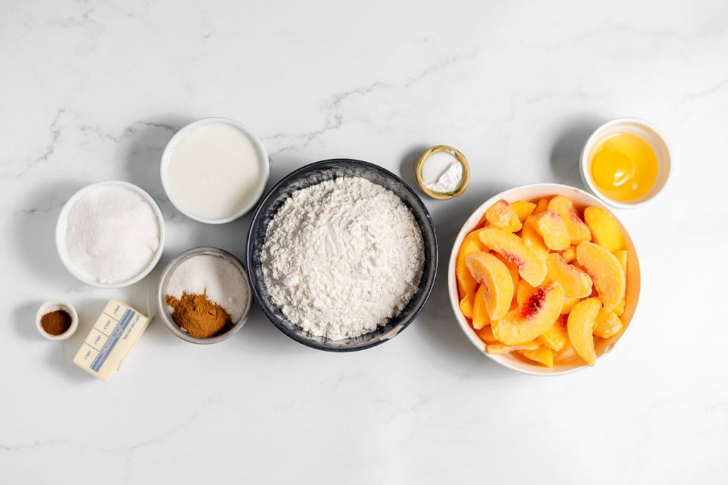 Ingredients for baking are arranged on a marble countertop, including flour, butter, sugar, spices, sliced peaches, milk, an egg, and baking powder.