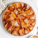 A round peach cake placed on a marble surface, with a bowl of fresh peach slices and a striped cloth nearby.