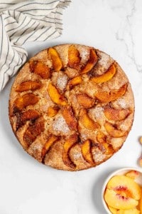 A round peach cake placed on a marble surface, with a bowl of fresh peach slices and a striped cloth nearby.