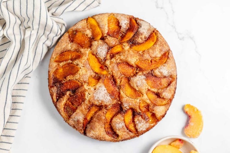 A round peach cobbler on a white marble surface, accompanied by a striped cloth and a small bowl with peach slices.