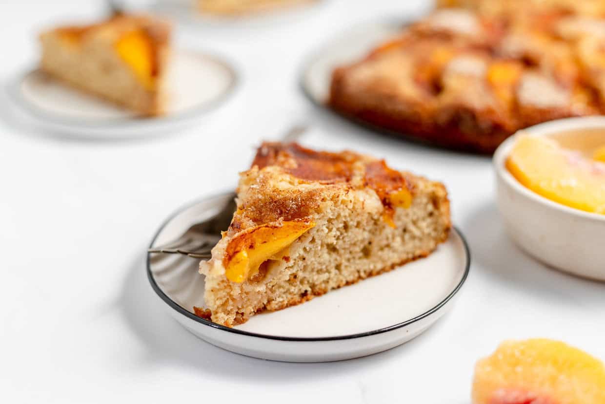 A slice of peach cake on a white plate with a fork, alongside another slice and a partial view of the remaining cake in the background.