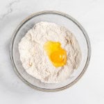 A clear glass bowl contains flour with a cracked egg yolk on top, set on a white marble surface.