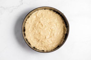 A round baking pan filled with raw cake batter, placed on a white marble surface.