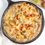 A cast iron skillet filled with a baked creamy dish topped with melted, browned cheese and garnished with chopped herbs, placed beside a cutting board with green bell pepper slices and mushrooms.