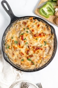 A cast iron skillet filled with a baked creamy dish topped with melted, browned cheese and garnished with chopped herbs, placed beside a cutting board with green bell pepper slices and mushrooms.