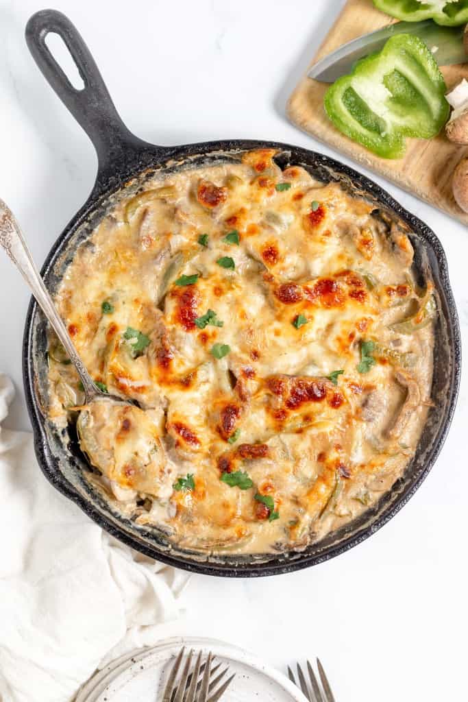 A cast iron skillet filled with baked creamy chicken and mushrooms, reminiscent of a Philly Cheesesteak Casserole, garnished with herbs, placed next to a white cloth, a fork, and a wooden board with bell pepper and mushrooms.