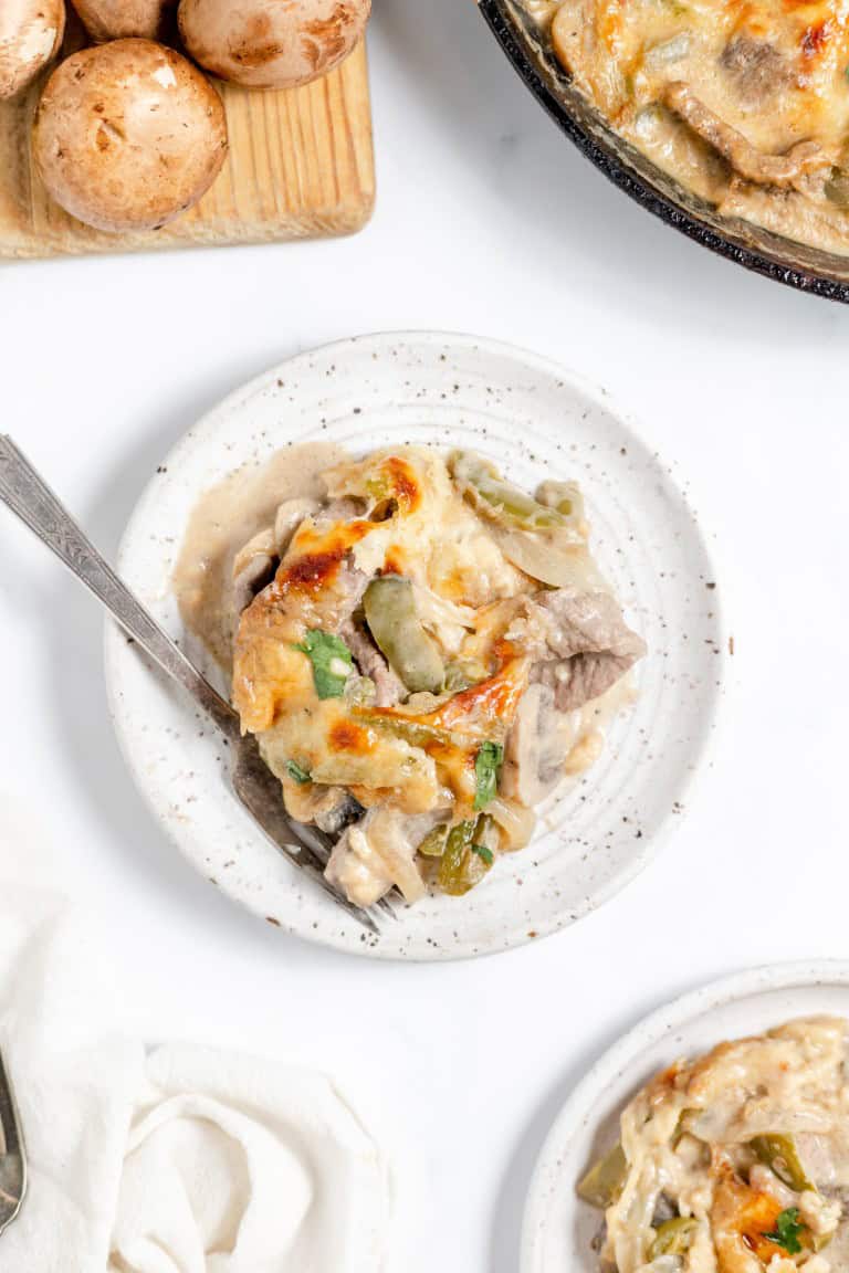 A plate of baked casserole with mushrooms and green vegetables next to a chopping board with whole mushrooms. A fork rests on the plate.