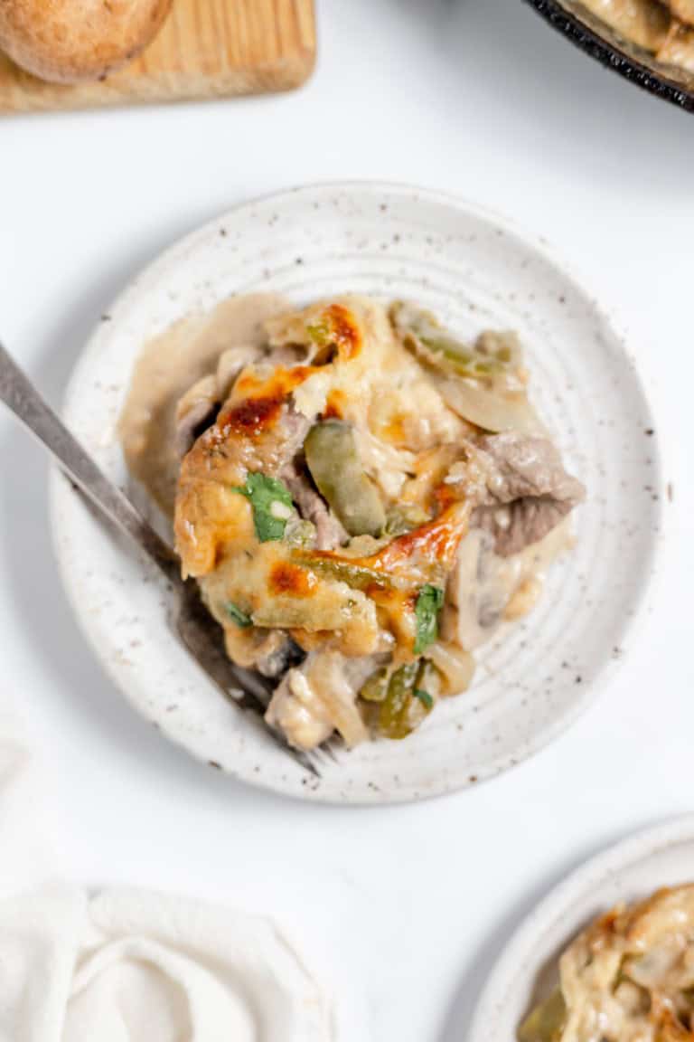 A cast iron skillet filled with baked creamy chicken and mushrooms, reminiscent of a Philly Cheesesteak Casserole, garnished with herbs, placed next to a white cloth, a fork, and a wooden board with bell pepper and mushrooms.