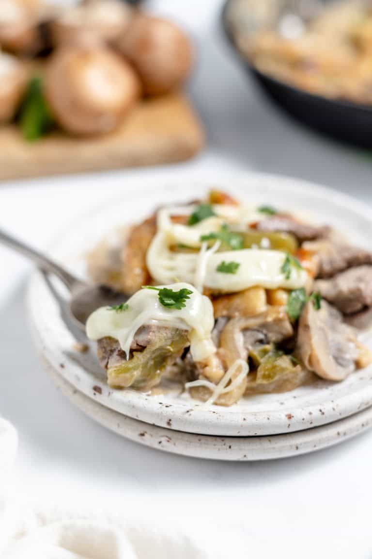 A plate of food featuring sliced mushrooms, melted cheese, and possibly meat, garnished with fresh cilantro. A fork rests on the plate, and a cutting board with whole mushrooms is visible in the background.