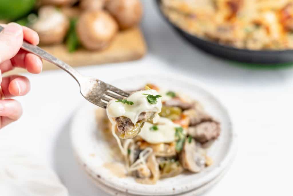 A close-up of a fork holding a bite of cheesy, mushroom-topped food over a plate, with a slice of a skillet dish and fresh mushrooms in the background.