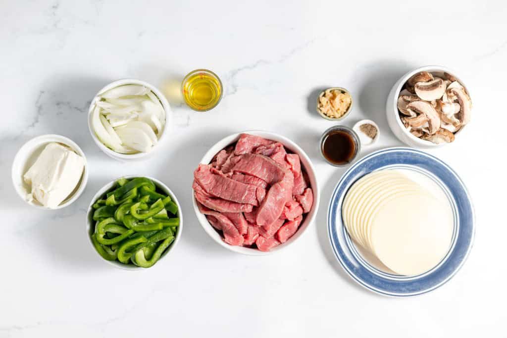 Ingredients for a Philly Cheesesteak Casserole arranged on a white surface: sliced onions, green bell peppers, raw beef strips, mushrooms, cheese slices, olive oil, minced garlic, ground pepper, and a small bowl of sauce.