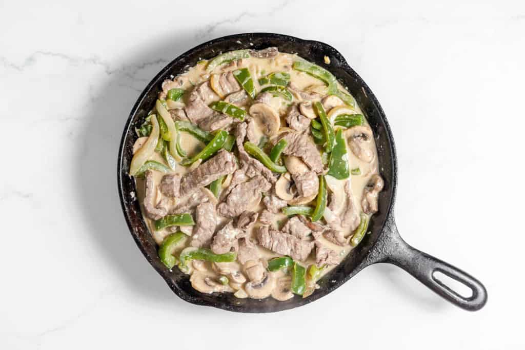 A cast iron skillet filled with a Philly Cheesesteak Casserole, featuring strips of beef, green bell peppers, and sliced mushrooms in a creamy sauce, placed on a white marble surface.