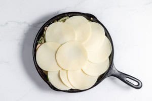 A cast iron skillet filled with Philly Cheesesteak Casserole ingredients, topped with a layer of circular slices of cheese, rests on a white marble surface.
