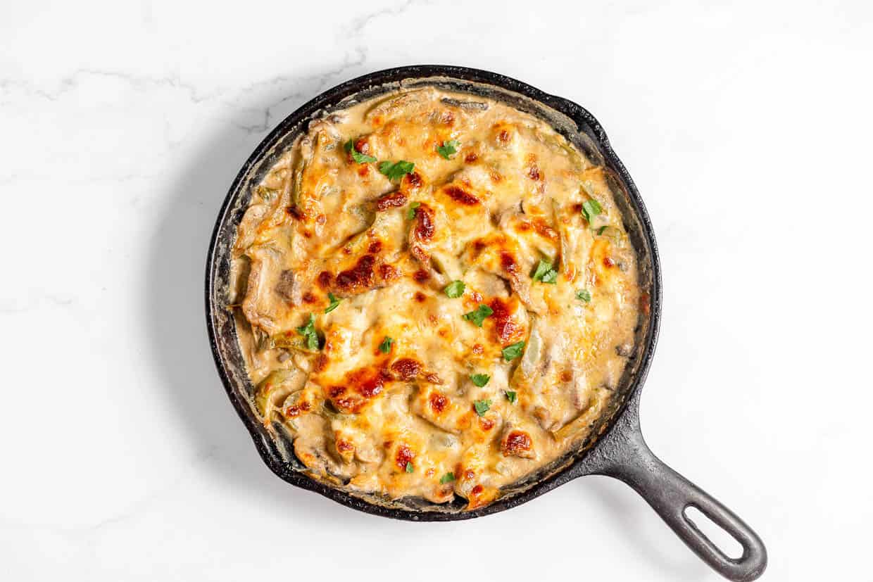 An overhead view of a cast iron skillet filled with a baked cheesy Philly Cheesesteak Casserole dish topped with chopped herbs, resting on a white marble surface.