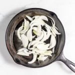 Sliced onions frying in a black cast iron skillet on a white marble surface create the perfect aroma for your upcoming Philly Cheesesteak Casserole.