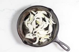 Sliced onions frying in a black cast iron skillet on a white marble surface create the perfect aroma for your upcoming Philly Cheesesteak Casserole.