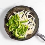 A cast iron skillet on a white surface contains sliced green bell peppers and onions, perfect ingredients for a Philly Cheesesteak Casserole.