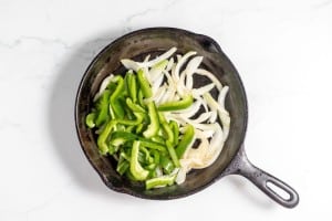 A cast iron skillet on a white surface contains sliced green bell peppers and onions, perfect ingredients for a Philly Cheesesteak Casserole.