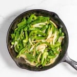 A cast iron skillet with saut&eacute;ed green bell peppers and onions, reminiscent of a Philly Cheesesteak Casserole, sits invitingly on a white surface.