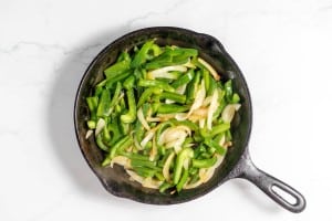 A cast iron skillet with sautéed green bell peppers and onions, reminiscent of a Philly Cheesesteak Casserole, sits invitingly on a white surface.
