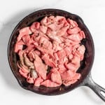 A cast iron skillet filled with raw, thinly sliced beef on a white marble countertop, ready to be transformed into a delicious Philly Cheesesteak Casserole.