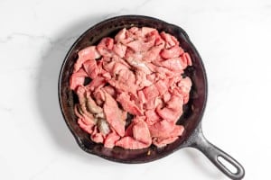 A cast iron skillet filled with raw, thinly sliced beef on a white marble countertop, ready to be transformed into a delicious Philly Cheesesteak Casserole.