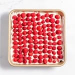 A symmetrical arrangement of raspberries on a square cake in a beige baking pan on a white marble surface.