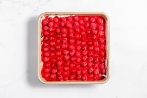 Square baking dish filled with cherry-topped dessert featuring a shiny red glaze, placed on a white surface.