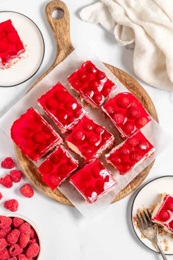 Top view of nine raspberry bars on a wooden serving board, with fresh raspberries in a bowl and two plates with raspberry bars, one partially eaten, on a white countertop.